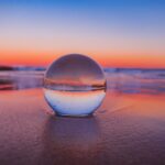 A glass ball sits on the beach at sunset.
